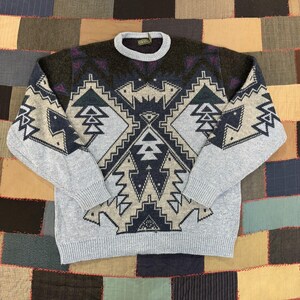 May include: A long-sleeve knit sweater featuring a crew neckline and a geometric pattern in shades of light blue, dark blue, charcoal gray, and purple. The design includes symmetrical angular shapes across the chest and sleeves with ribbed cuffs.