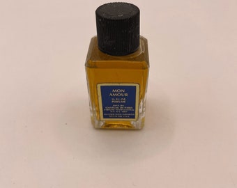 May include: A small, rectangular glass perfume bottle filled with amber-colored liquid, topped with a black ribbed cap. The blue label reads MON AMOUR, 1/2 FL. OZ. PERFUME, DIST. BY CHATEAU DE PARIS PARFUME INTERNATIONAL N.Y., N.Y. 10017.