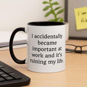 Op de afbeelding: Witte keramische mok met een zwarte binnenkant en handvat. De mok heeft de tekst: "I accidentally became important at work and it's ruining my life."