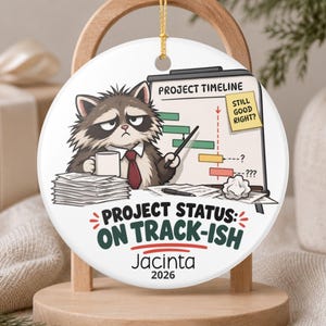 May include: A round white ceramic ornament featuring a cartoon raccoon in a tie holding a mug, pointing at a project timeline chart with a sticky note saying STILL GOOD RIGHT? Text reads PROJECT STATUS: ON TRACK-ISH, Jacinta, and 2026.