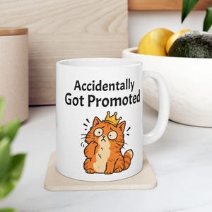 May include: A white ceramic mug featuring the black text Accidentally Got Promoted above a cartoon illustration of an orange tabby cat wearing a small gold crown with a surprised and concerned facial expression.