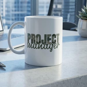 May include: White ceramic mug with the words "PROJECT manager" printed in a camouflage and script font. The mug has a curved handle and is sitting on a light gray surface. A laptop and pen are visible in the background.