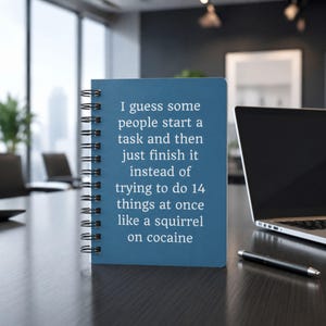 May include: A blue spiral-bound notebook featuring white text that reads: I guess some people start a task and then just finish it instead of trying to do 14 things at once like a squirrel on cocaine. The notebook has a black metal coil binding.
