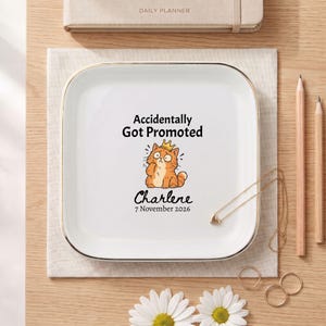 Puede incluir: Un plato cuadrado de cerámica blanca con un borde dorado. El centro presenta un gráfico de un gato naranja con una pequeña corona dorada, junto con el texto "Accidentally Got Promoted", el nombre "Charlene" y la fecha "7 November 2026".