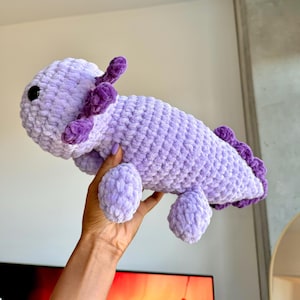 Puede incluir: Un peluche de axolotl lavanda con branquias, crestas de la cabeza y aleta caudal de color púrpura oscuro. El animal de peluche hecho a mano tiene pequeños ojos negros y pies redondeados. El material suave y texturizado le da al juguete una apariencia tierna.