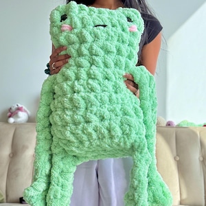May include: A large, light green plush frog made of chunky, textured yarn. The frog features two dark eyes, a small horizontal line for a mouth, and two small pink circles on its cheeks. It has long, dangling arms and legs with a soft, fuzzy appearance.