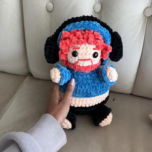 Puede incluir: Muñeca de ganchillo hecha a mano con gorro y suéter azules, pantalón negro y barba roja. La muñeca tiene ojos negros y cara y manos color melocotón. La muñeca está sentada.