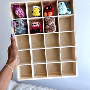May include: A light-colored wooden display shelf with twenty square compartments. Seven small, plush, crocheted animal figures in various colors including pink, yellow, red, blue, gray, and brown are arranged in the top two rows of the grid.