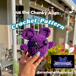 Könnte beinhalten: Ein lila gehäkeltes Alien-Spielzeug mit großen schwarzen Augen, kleinen Armen und Ohren. Der Text "Gus the Chonky Alien" und "Crochet Pattern" sind über dem Spielzeug zu sehen. Das Spielzeug wird in einer Hand gehalten, mit dem Logo @crochetwithashroot und Arthyria unten rechts.