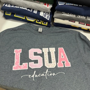 Könnte beinhalten: Ein dunkelgrau meliertes Kurzarm-T-Shirt mit den Buchstaben LSUA als Applikation aus rosa-weiß gemustertem Stoff. Unter den Buchstaben ist das Wort education in weißer Schreibschrift gedruckt. Ein Stapel ähnlicher T-Shirts ist im Hintergrund zu sehen.
