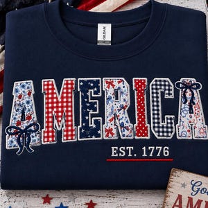 May include: A navy blue crewneck sweatshirt featuring the word AMERICA in large, patterned fabric letters with red, white, and blue designs. The text EST. 1776 is printed in white below the word, underlined by a thin red line. The shirt has a Gildan label.