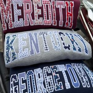 May include: Three sweatshirts stacked on top of each other. The top sweatshirt is maroon with the name "MEREDITH" in white outlined letters with floral and gingham patterns. The middle sweatshirt is gray with "KENTUCKY" in white outlined letters with floral and striped patterns. The bottom sweatshirt is navy blue with "GEORGETOWN" in white outlined letters with floral, gingham, and polka dot patterns.