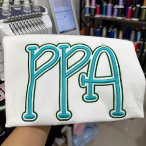 May include: White fabric with the embroidered letters "PPA" in teal, outlined in green. The letters have a rounded, playful font. The fabric is held up in front of an embroidery machine and spools of thread.