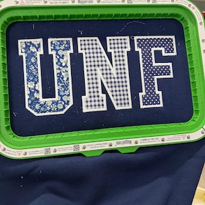 May include: A navy blue fabric is held in a green embroidery hoop. The letters "UNF" are embroidered on the fabric. The "U" is floral, the "N" is checkered, and the "F" is polka-dotted. A Brother sewing machine is visible in the background.