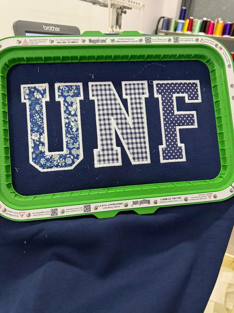 May include: A navy blue fabric is held in a green embroidery hoop. The letters "UNF" are embroidered on the fabric. The "U" is floral, the "N" is checkered, and the "F" is polka-dotted. A Brother sewing machine is visible in the background.