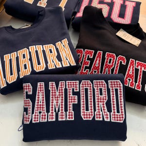 Op de afbeelding: Een verzameling marineblauwe en zwarte sweatshirts met college-letters. De sweatshirts tonen de namen "AUBURN", "BEARCAT", "SAMFORD" en "FSU" in verschillende kleuren en patronen, waaronder oranje strepen en rood ruitjespatroon.