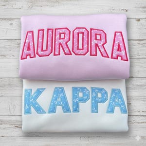 Op de afbeelding: Twee sweatshirts, één roze en één wit, op elkaar gestapeld. Het roze sweatshirt heeft de naam "AURORA" geborduurd in roze met een bloemenpatroon. Het witte sweatshirt heeft "KAPPA" geborduurd in blauw met een bloemenpatroon.
