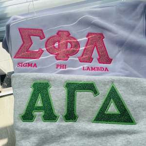 Op de afbeelding: Er worden twee sweatshirts getoond, een lichtpaarse en een grijze. Het lichtpaarse sweatshirt heeft roze glitterletters met de tekst "SIGMA PHI LAMBDA". Het grijze sweatshirt heeft groene glitterletters met de tekst "ALPHA GAMMA".