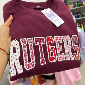 Pode incluir: Uma camisola bordô com a palavra "RUTGERS" em letras grandes e decorativas. As letras apresentam uma mistura de padrões, incluindo bolinhas, estampas florais e xadrez. Uma etiqueta com o nome da marca está anexada.