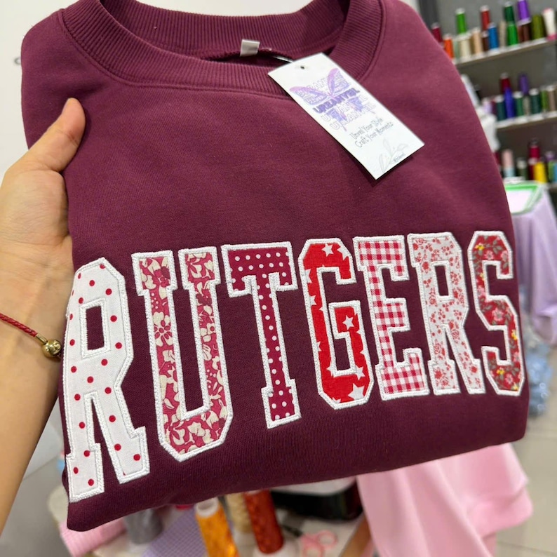 May include: A maroon sweatshirt with the word "RUTGERS" in large, decorative letters. The letters feature a mix of patterns, including polka dots, floral prints, and gingham. A tag with the brand name is attached.