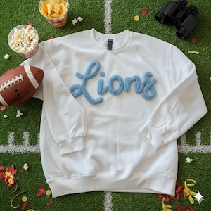 Könnte beinhalten: Weißes Sweatshirt mit dem Wort "Lions" in hellblauen, flauschigen Buchstaben. Das Sweatshirt liegt auf einer grünen Oberfläche mit einem Football, Popcorn, Chips und einem Fernglas.