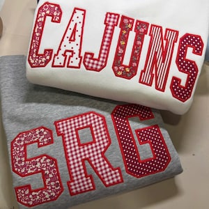May include: Two sweatshirts, one white and one gray, are stacked. The white sweatshirt has the word "CAJUNS" in red, patterned lettering. The gray sweatshirt displays the letters "SRG" in a similar red, patterned style. The letters are outlined in red.