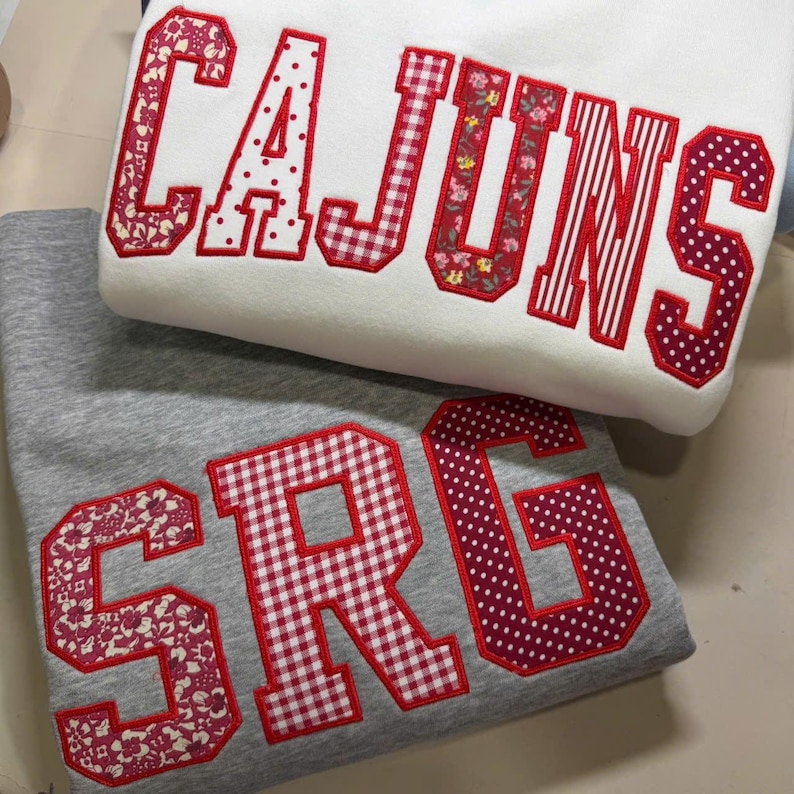 May include: Two sweatshirts, one white and one gray, are stacked. The white sweatshirt has the word "CAJUNS" in red, patterned lettering. The gray sweatshirt displays the letters "SRG" in a similar red, patterned style. The letters are outlined in red.