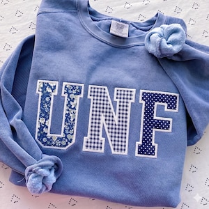 May include: A light blue sweatshirt with the letters "UNF" in a floral, gingham, and polka dot pattern. A matching scrunchie rests on the shoulder. The tag reads "Comfort Colors".
