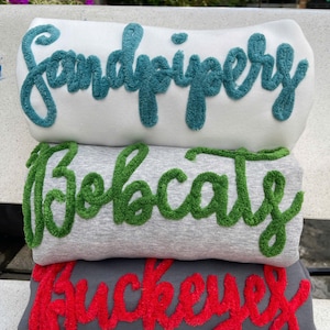 Könnte beinhalten: Drei gefaltete Sweatshirts übereinander gestapelt. Das obere Sweatshirt ist weiß mit dem Wort "Sandpiper" in türkisfarbener Schrift. Das mittlere Sweatshirt ist grau mit "Bobcats" in grüner Schrift. Das untere Sweatshirt ist dunkelgrau mit "Buckeyes" in roter Schrift.
