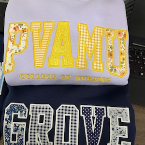 May include: Two sweatshirts are stacked, one lavender and one navy blue. The lavender sweatshirt has the text "PVAMU COLLEGE OF NURSING" in yellow and floral patterns. The navy blue sweatshirt displays the word "GROVE" in various patterns.