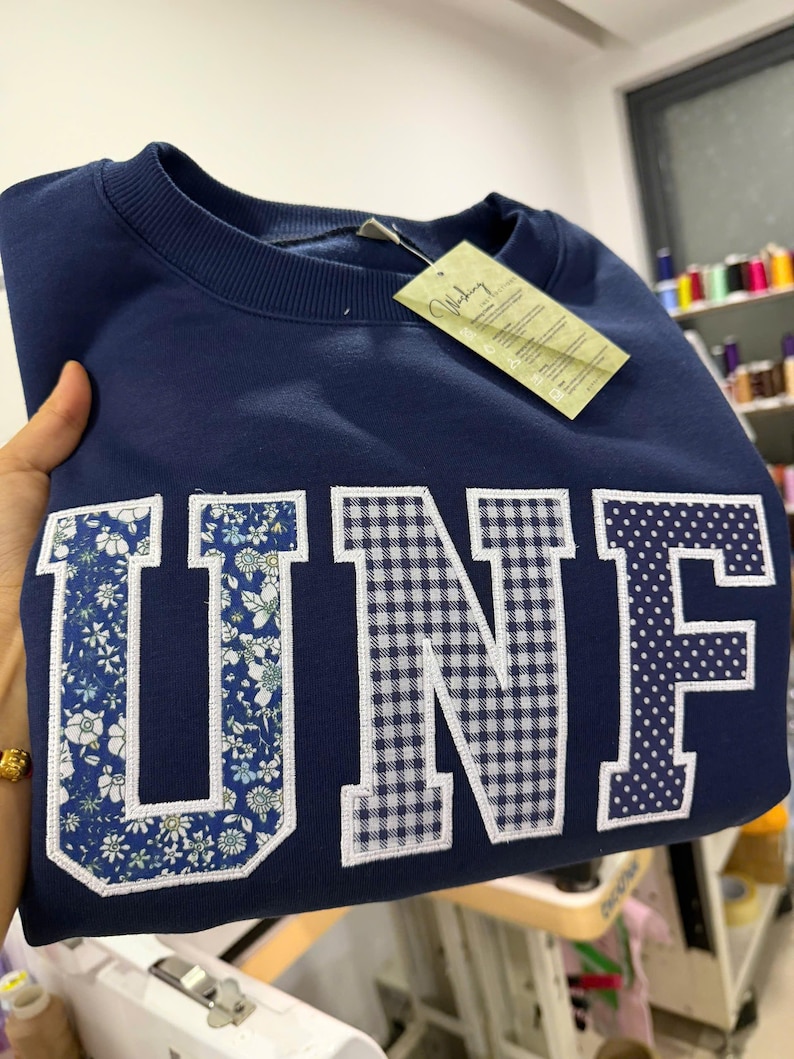 May include: Navy blue sweatshirt with the letters "UNF" in a floral, gingham, and polka dot design. The sweatshirt has a crew neck and a tag attached. The letters are outlined in white.