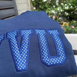 Puede incluir: Sudadera azul marino con las letras "VU" en tela de lunares blancos. Las letras est&aacute;n delineadas en azul marino y cosidas a la sudadera. El fondo es azul marino liso.