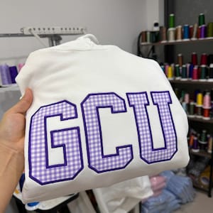 May include: A white hooded sweatshirt featuring the letters GCU in large, block-style applique. The letters are filled with a purple and white gingham check pattern and are outlined with a thick, dark purple embroidered border.
