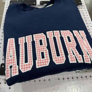 May include: A navy blue crewneck sweatshirt featuring the word AUBURN in large, block-style letters. The letters are filled with a pink and white gingham check pattern and are outlined with white stitching against the dark blue fabric background.