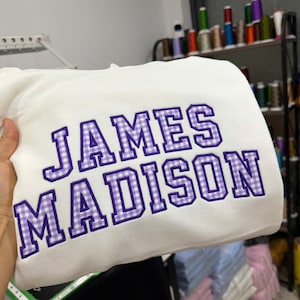 May include: A white sweatshirt featuring the embroidered text JAMES MADISON in a collegiate font. The letters are filled with a purple and white gingham check pattern and outlined in a dark purple thread, creating a bold, textured appearance.