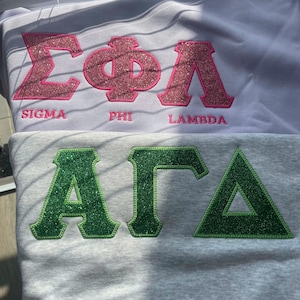 Op de afbeelding: Twee sweatshirts, één witte en één grijze, elk met Griekse letters. Het witte sweatshirt heeft roze glitterletters met de tekst "Sigma Phi Lambda" met de woorden "Sigma", "Phi" en "Lambda" eronder geborduurd. Het grijze sweatshirt heeft groene glitterletters.