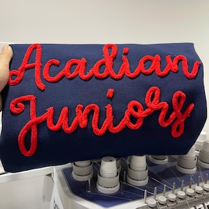 Peut inclure: Tissu bleu marine avec l'inscription "Acadian Juniors" brodée en rouge, en lettres cursives. Le tissu est tenu par une main, et l'arrière-plan montre une machine à coudre.
