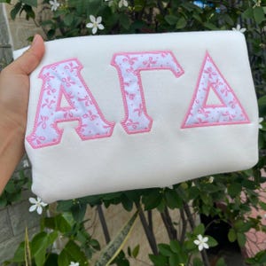 May include: A white sweatshirt featuring the Greek letters ΑΓΔ in a pink floral pattern with a pink border. The letters are large and prominently displayed on the front of the sweatshirt.