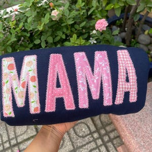 Op de afbeelding: Een marineblauw kussen met het woord "MAMA" in applicatiestof. De letters zijn in roze en bloemmotieven, afgezet met roze kant. Het kussen wordt vastgehouden voor een groene, bladerrijke achtergrond.