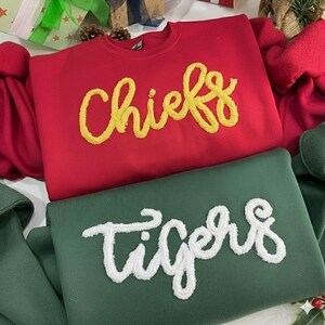 May include: Two sweatshirts, one red and one green. The red sweatshirt has the word "Chiefs" in yellow script. The green sweatshirt has the word "Tigers" in white script. Both sweatshirts are made of a soft material.