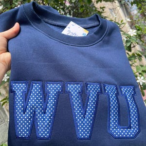 Könnte beinhalten: Marineblaues Sweatshirt mit den Buchstaben "WVU" in einem weißen Polka-Dot-Muster. Die Buchstaben sind dunkelblau umrandet. Das Sweatshirt ist gefaltet.