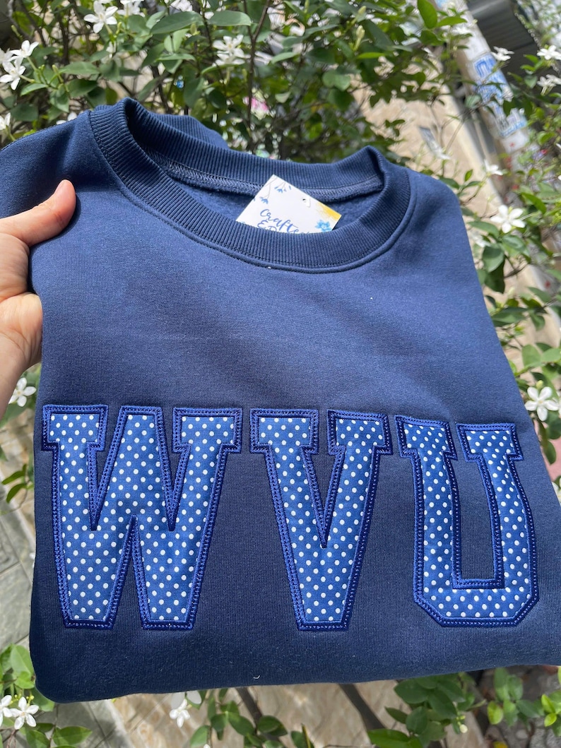 Puede incluir: Sudadera azul marino con las letras "WVU" en un patr&oacute;n de lunares blancos. Las letras est&aacute;n delineadas en azul m&aacute;s oscuro. La sudadera est&aacute; doblada.