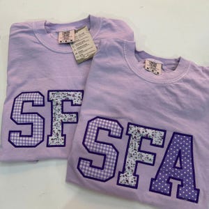 May include: Two lavender Comfort Colors crewneck sweatshirts. Each shirt features a large, appliqued letter design. One shirt has "SF" with gingham and floral patterns, the other has "SFA" with gingham and polka dot patterns. Both have tags.