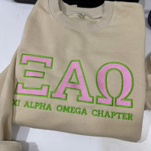 May include: A beige sweatshirt with the Greek letters ΕΑΩ in pink with green outlines. The text "XI ALPHA OMEGA CHAPTER" is embroidered in green below the letters. The sweatshirt has a crew neck and long sleeves.