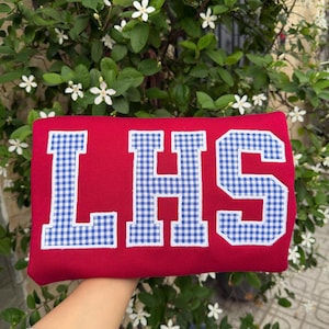 May include: A bright red sweatshirt featuring the letters LHS in a blue and white gingham check pattern. Each letter is outlined with white stitching, creating a bold, block-style appliqu&eacute; design centered on the front of the garment.