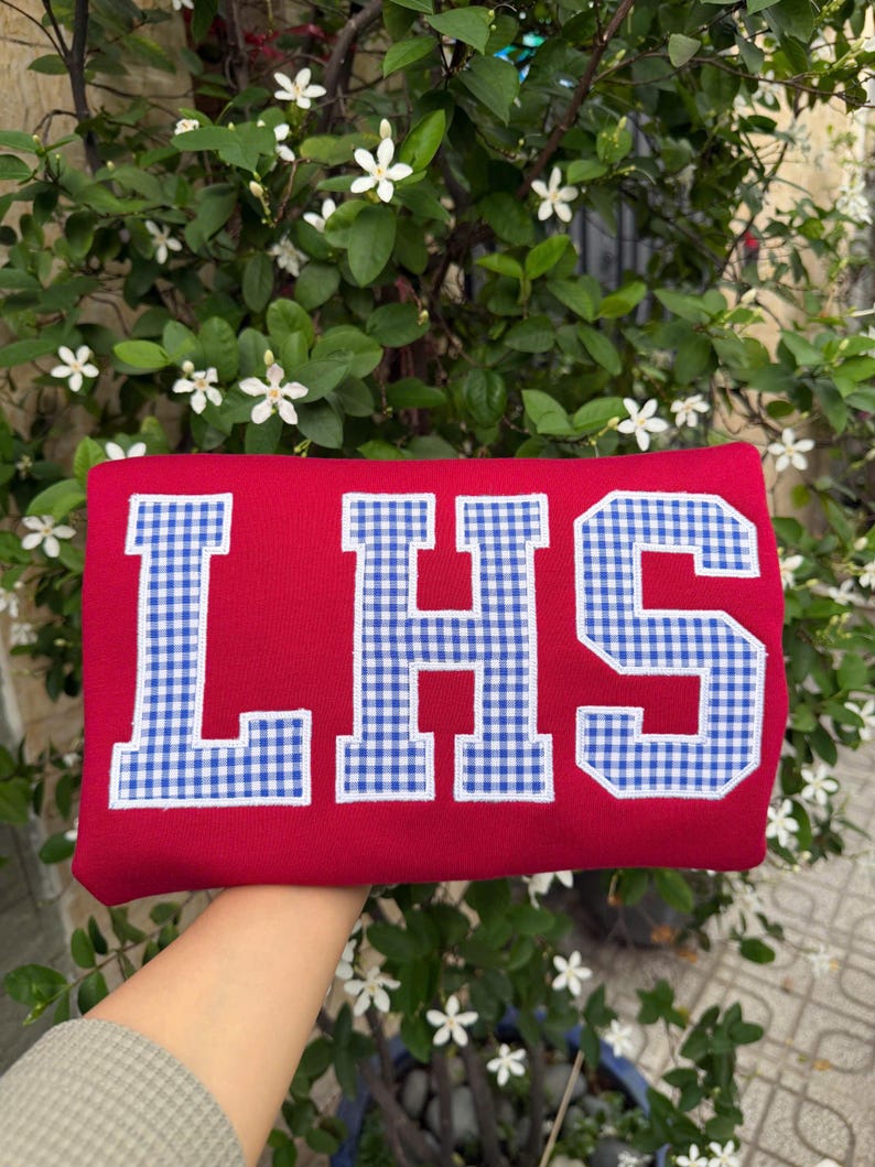 May include: A bright red sweatshirt featuring the letters LHS in a blue and white gingham check pattern. Each letter is outlined with white stitching, creating a bold, block-style appliqu&eacute; design centered on the front of the garment.