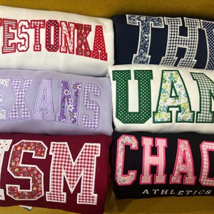 May include: Several folded sweatshirts in different colours, each with a word or name in large, decorative lettering. The lettering includes floral, polka dot, and gingham patterns. The sweatshirts are stacked on a yellow surface.