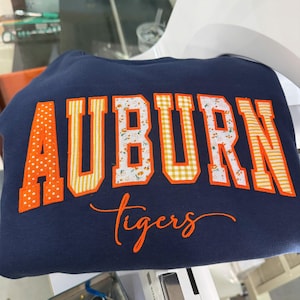 May include: A navy blue sweatshirt featuring the word AUBURN in large, block-style letters with orange and white patterned fabric appliques. Below the word AUBURN, the word tigers is embroidered in orange cursive script lettering.