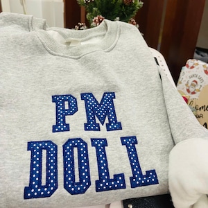 Może przedstawiać: Jasnoszara bluza z napisem "PM DOLL" w niebieskich literach w kropki. Bluza ma okrągły dekolt i długie rękawy. Mankiety są białe.