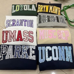 May include: A collection of folded sweatshirts in various colors, each embroidered with a different college or university name. The lettering features floral, plaid, and striped patterns. The sweatshirts are in shades of green, navy, white, and beige.